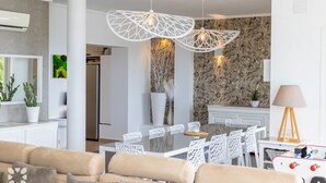 Un salón moderno con un sofá color beige, sillas blancas, una mesa de comedor y una zona de cocina con salpicadero de piedra.