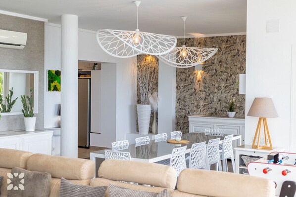 A modern living room with a beige sofa, white chairs, a dining table, and a kitchen area with a stone backsplash.