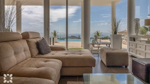 Un salón espacioso con un amplio sofá seccional color beige, una mesa de centro de vidrio y vistas a una piscina y montañas.