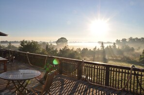A wooden deck with a round table and chairs, overlooking a misty landscape with trees and a body of water. - Bright, cheerful apartment with piano in the heart of East Friesland (Großefehn)