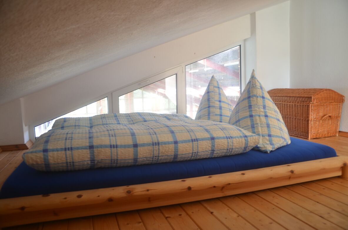 A loft bedroom with a sloped ceiling, a blue mattress, plaid pillows, a wicker basket, and large windows.