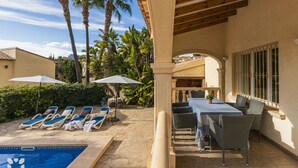 A pool area with lounge chairs and umbrellas, a covered patio with a table and chairs, and a view of palm trees and a mountain in the background.