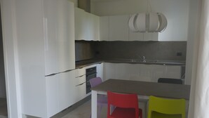 Moderni keittiö, jossa on valkoiset kaapit, harmaa työtaso ja riippuvalaisin. Keittiössä on kaksi tuolia, toinen punainen ja toinen keltainen, sekä yksi violetti tuoli.
