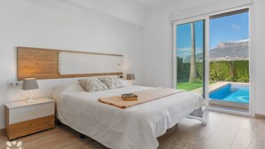 A bedroom with a large bed, a nightstand, a lamp, and a view of a swimming pool through a sliding glass door. - Villa SOCRATES by Abahana Villas (Calpe)