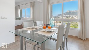 A dining area with a glass table, white chairs, and a breakfast setting including orange juice and fruits. - Villa SOCRATES by Abahana Villas (Calpe)