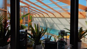 Un espace piscine intérieur doté d’un plafond en bois et de panneaux de verre, entouré de plantes en pot et agrémenté d’un fauteuil de relaxation.
