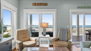 Una sala de estar con dos sillones color beige, una mesa de centro blanca y vista a la playa a través de amplias ventanas.