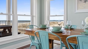 Un comedor con mesa de madera y sillas azul verdosas, ventanas amplias con vista a la playa, y un cuadro en la pared.