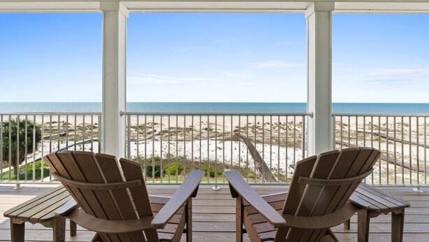 Una terraza techada con dos sillas Adirondack y una mesa de madera, con vista a una playa de arena y un cielo azul despejado.