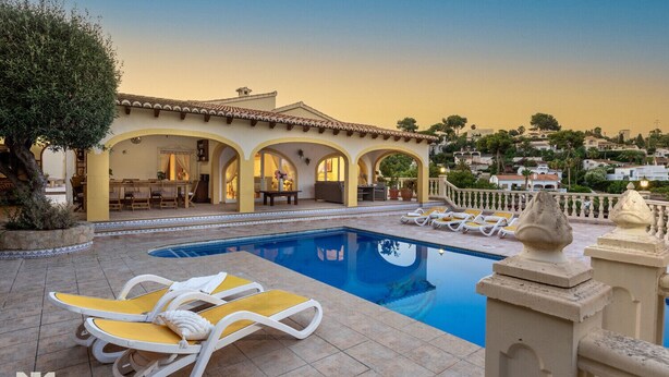 Un área de piscina con sillones, un comedor con mesas y sillas, y un edificio con arcos y tejado de azulejos.