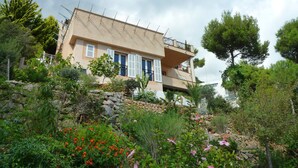Ein zweistöckiges Haus mit einer Stein-Fassade, einem Balkon im oberen Geschoss und einem Garten mit verschiedenen Pflanzen und Blumen.