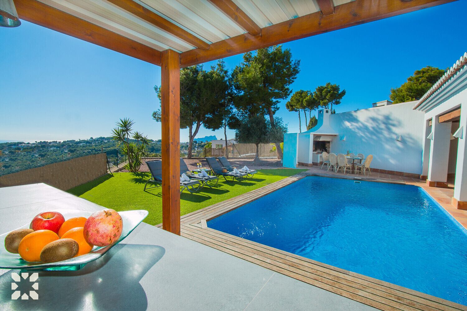 Une piscine avec une terrasse en bois, une table garnie de fruits et des sièges d’extérieur.