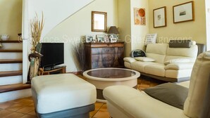 Een woonkamer met een bank, een poef en een salontafel. Er is een televisie op een meubel, een open haard en een trap.