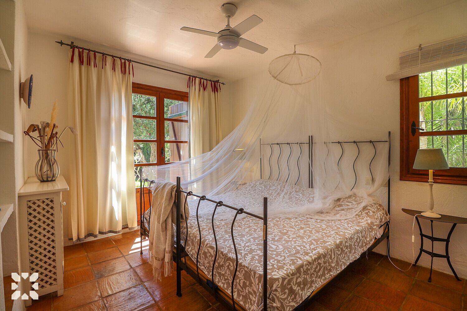 A bedroom with a bed, a ceiling fan, a window with curtains, a bedside table with a lamp, and a decorative vase.