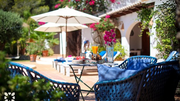 Un espacio de estar al aire libre con sillas azules, una mesa con un vaso de jugo de naranja, un florero con flores rosadas y un paraguas blanco.