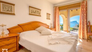 Une chambre avec un grand lit, une tête de lit en bois et une table de chevet avec une lampe. Deux tableaux encadrés ornent le mur, et la fenêtre offre une vue sur la mer.
