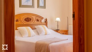 Un lit bien fait, avec une tête de lit en bois, des oreillers blancs et une couverture beige. Une table de chevet avec une lampe se trouve à côté du lit, et un tableau encadré est accroché au mur au-dessus du lit.