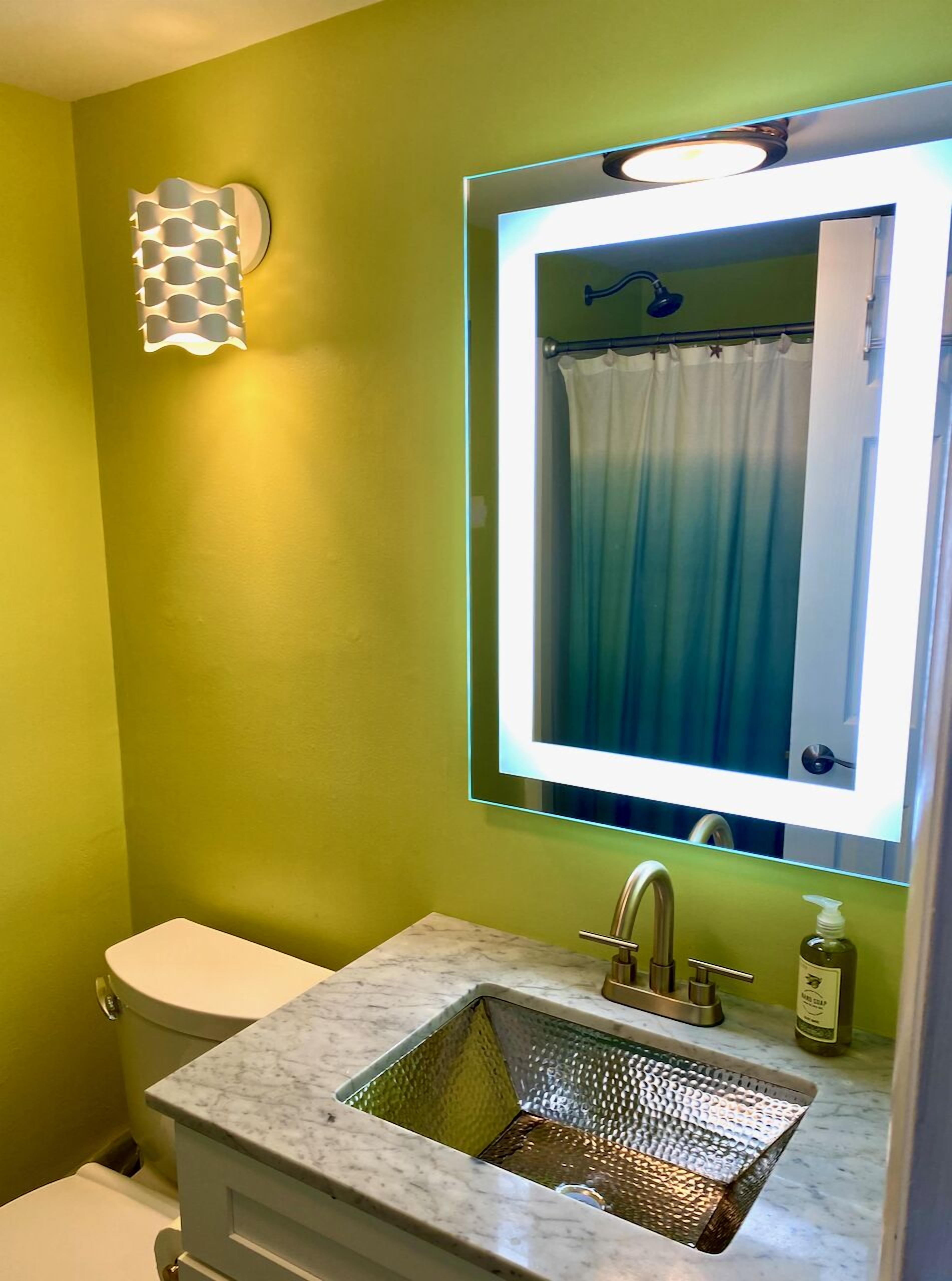 A bathroom with a green wall, a white toilet, a sink with a marble countertop, and a mirror with a light.