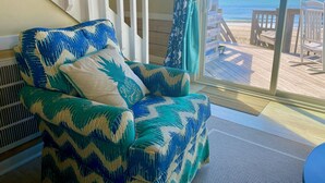 A room with a view of the beach, a chair with a patterned cover, and a window with curtains. - Oceanfront Luxury House with Stunning Views (Emerald Isle)