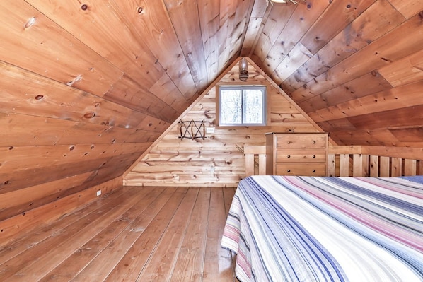 Une chambre mansardée en bois, avec un plafond en pente, une fenêtre, un lit et une commode.
