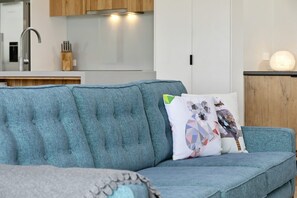 A modern living room with a blue sofa, a decorative pillow, and a kitchen in the background.