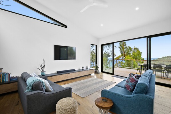 A modern living room with a flat-screen TV, two sofas, a coffee table, and a view of the outdoors through large windows.