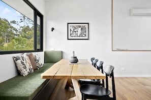 Een eetgedeelte met een houten tafel, zwarte stoelen, een bank met kussens, een ingelijst kunstwerk en een raam met uitzicht op groen.