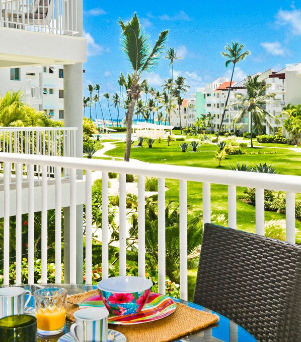 A balcony with a table set for breakfast, overlooking a well-maintained garden and a pool.