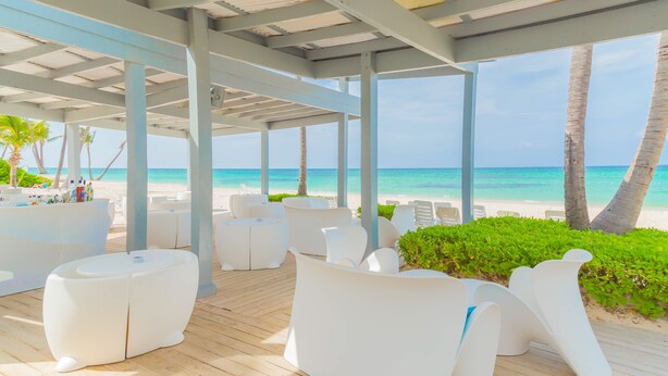 Un espace extérieur aménagé, abrité et doté de mobilier blanc, d’une terrasse en bois et offrant une vue sur la plage et l’océan.