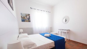 Quarto de hotel com cama, mesa de cabeceira com abajur, pequena escrivaninha, espelho e um quadro na parede.