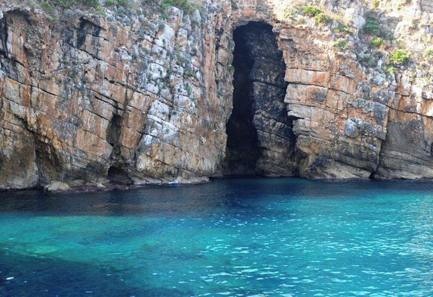 A rocky coastal cliff with a natural cave entrance and clear blue water.