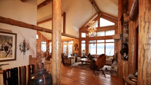 A spacious room with wooden beams, a large chandelier, and a view of the outdoors. - Sunset Cabin - Flathead Lake, Exquisite Log Home and Guest Cabin. Sleeps 12 (Bigfork)