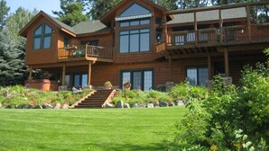 A large wooden cabin with a balcony, a staircase leading to the entrance, and a well-maintained garden. - Sunset Cabin - Flathead Lake, Exquisite Log Home and Guest Cabin. Sleeps 12 (Bigfork)