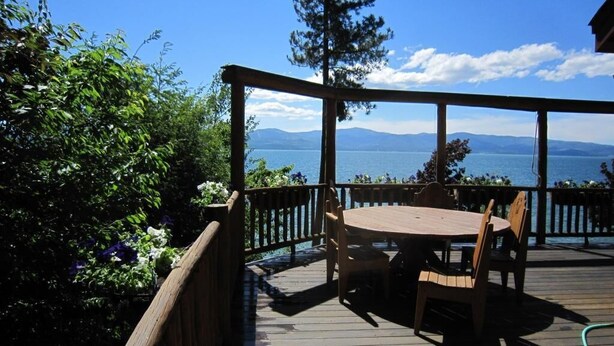 A wooden deck with a table and chairs, overlooking a body of water and mountains. - Sunset Cabin - Flathead Lake, Exquisite Log Home and Guest Cabin. Sleeps 12 (Bigfork)
