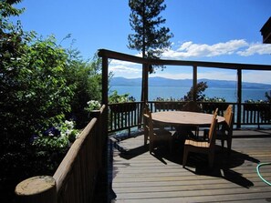 Um deck de madeira com mesa e cadeiras, com vista para um corpo d'água e montanhas.