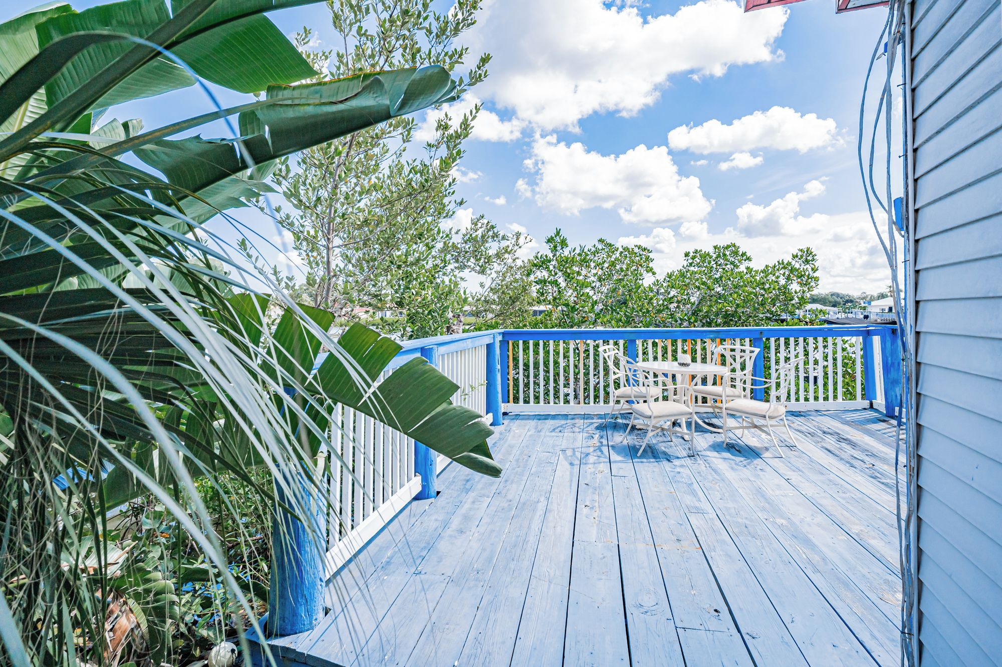 Eine Holzterrasse mit blauer Geländer, weißen Stühlen und Blick auf Bäume und Himmel.