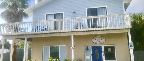 A two-story building with a blue exterior, a balcony with white railings, and a blue door with a number plaque.