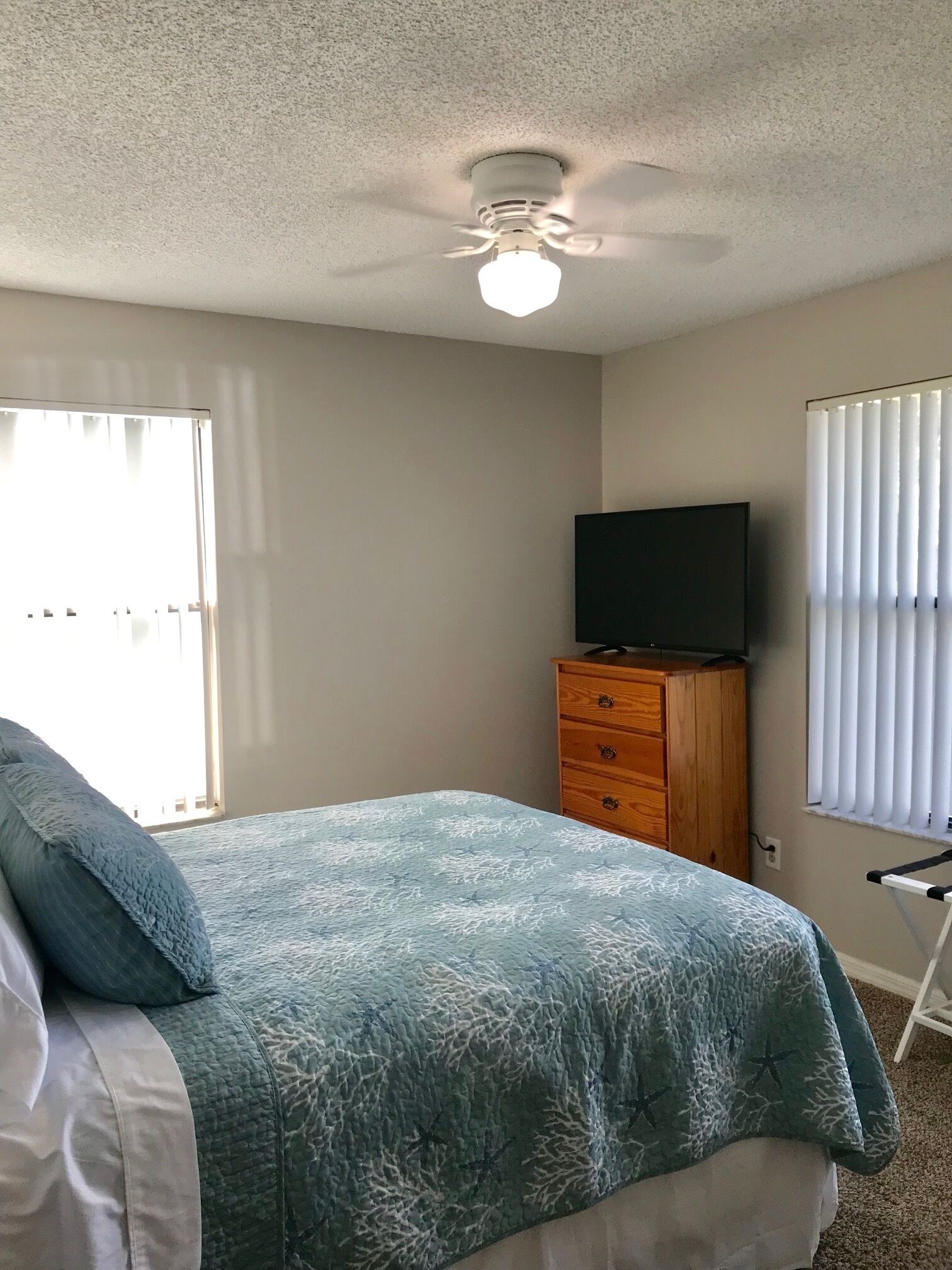 Un dormitorio con cama, ventilador de techo, televisor sobre una cómoda de madera y ventana con persianas.