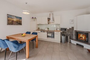 Une cuisine moderne avec des meubles blancs, un coin repas avec une table en bois et des chaises bleues, et une cheminée avec un feu visible.