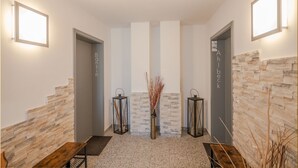 Uma área de recepção com parede de pedra, duas portas identificadas como “Banshin” e “Ahlbeck”, e um vaso central com plantas secas.