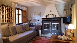 A cozy living room with a sofa, a fireplace, a wooden table with glassware, and a wall-mounted shelf with jars. - Charming country house in a quiet location with fantastic views and pool (Aramo)
