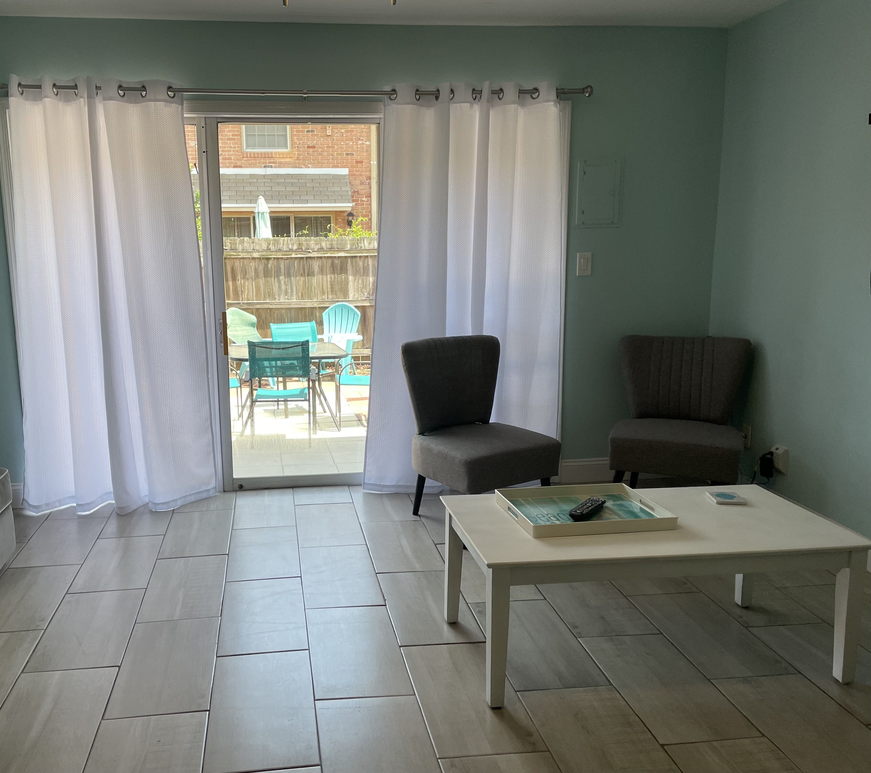 A room with a sliding glass door leading to an outdoor patio, two chairs, a table with a remote control and a book, and a light-colored floor.