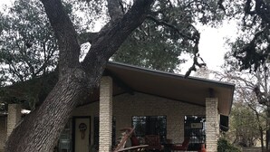 Un edificio rústico con fachada de piedra, un porche con sillas rojas y un gran árbol en primer plano.