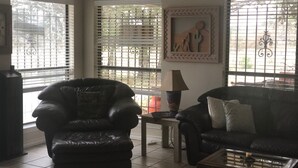 Una sala de estar con un sillón de cuero negro, un sofá negro y una mesa de centro. Hay dos ventanas con persianas, una lámpara sobre una mesita auxiliar y un cuadro en la pared.