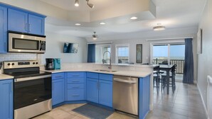 A modern kitchen with blue cabinets, stainless steel appliances, and a dining area with a view. - Love at First Flight: Oceanfront, Shared Pool (Kill Devil Hills)