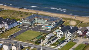 An aerial view of a coastal residential area with a large building complex, a beach, and a road. - Love at First Flight: Oceanfront, Shared Pool (Kill Devil Hills)