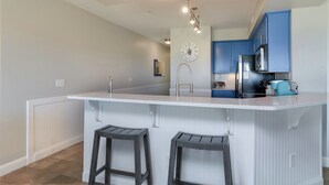 A modern kitchen with a central island, bar stools, and blue cabinets. - Love at First Flight: Oceanfront, Shared Pool (Kill Devil Hills)