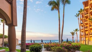 A resort courtyard with palm trees, a pool, and a view of the ocean. - Surf & Stay; Beachfront Condo Steps from Sand - Heated Hot tub -Pool-Pickleball (San José del Cabo)