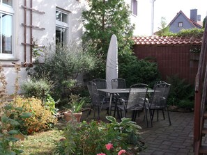 Um pátio externo com mesa e cadeiras, cercado por plantas em vasos e um cercado de madeira.