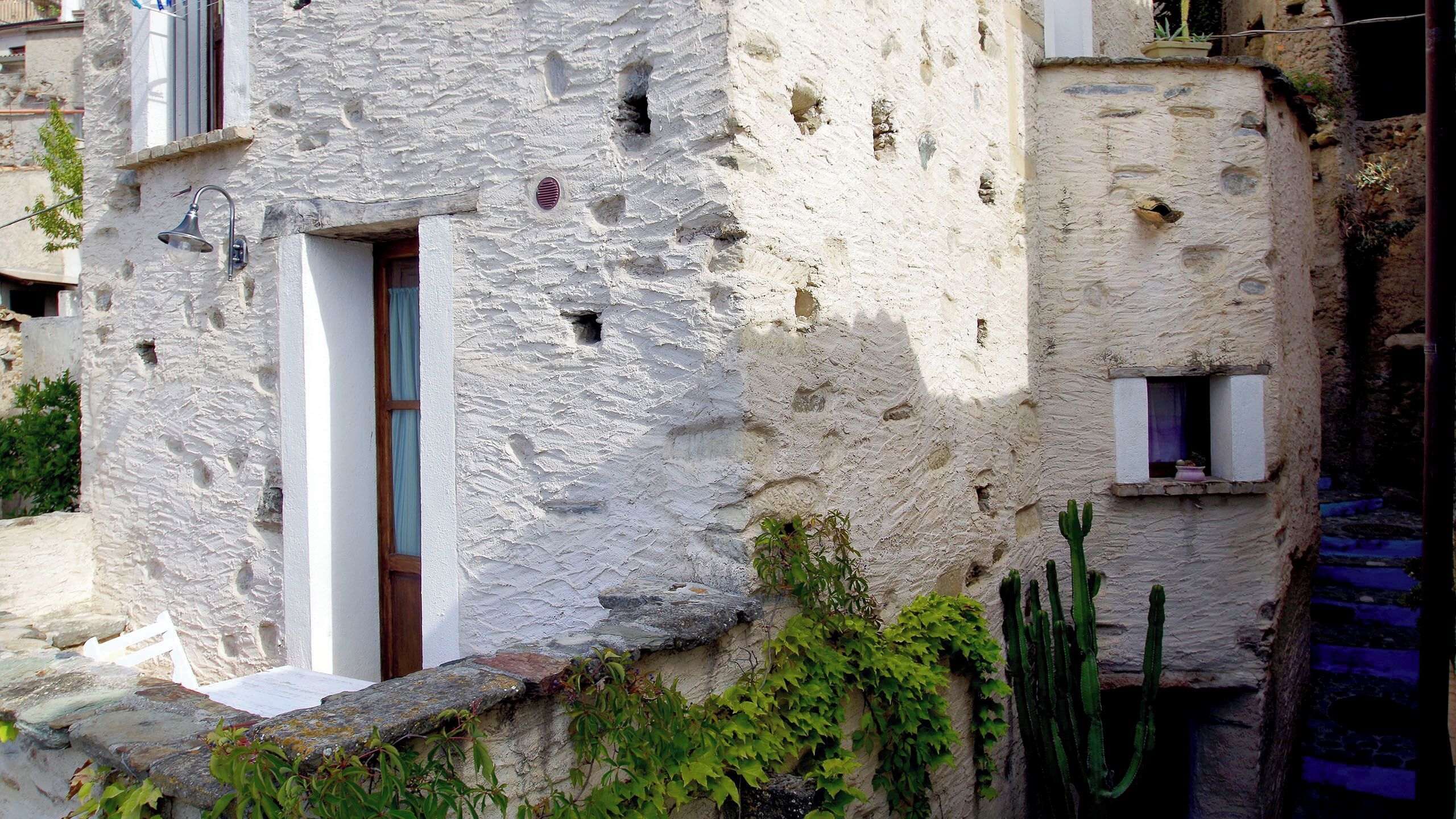 Un edificio de piedra tradicional con una ventana pequeña y una puerta, rodeado de follaje y un cactus.